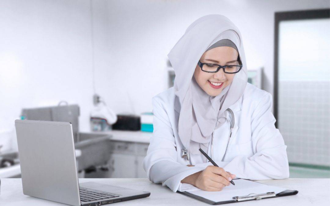 a female doctor is writing on a clipboard in front of a laptop